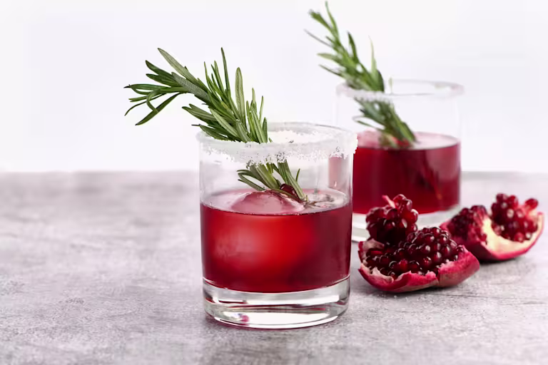 A glass of red cocktail with ice, garnished with a rosemary sprig and a sugared rim, sits on a gray surface with pomegranate seeds and another drink in the background.