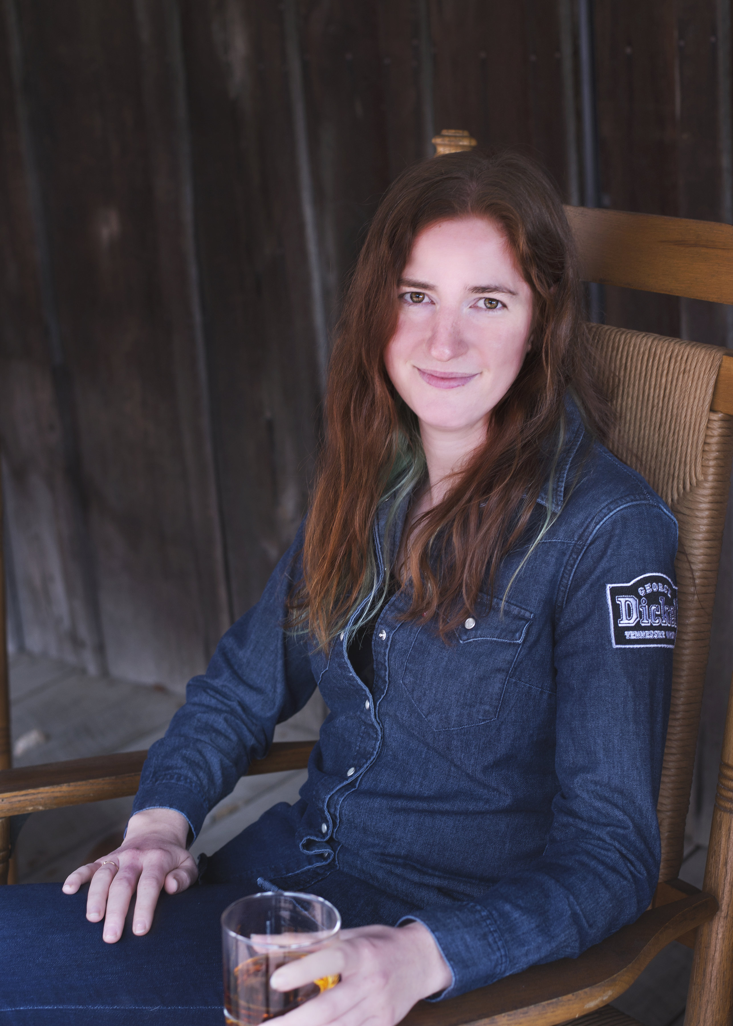 A woman with long brown hair sits in a wooden chair, wearing a blue denim shirt and holding a glass of beverage, with a dark wooden background.