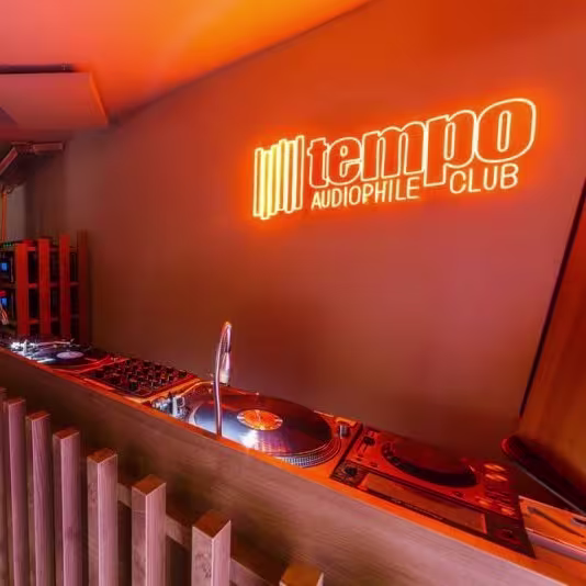 La habitación está iluminada con una luz ambiental cálida. A la izquierda, hay un mostrador de madera con taburetes delante, y detrás de este mostrador hay equipos de audio, incluyendo un amplificador y tocadiscos para vinilos..