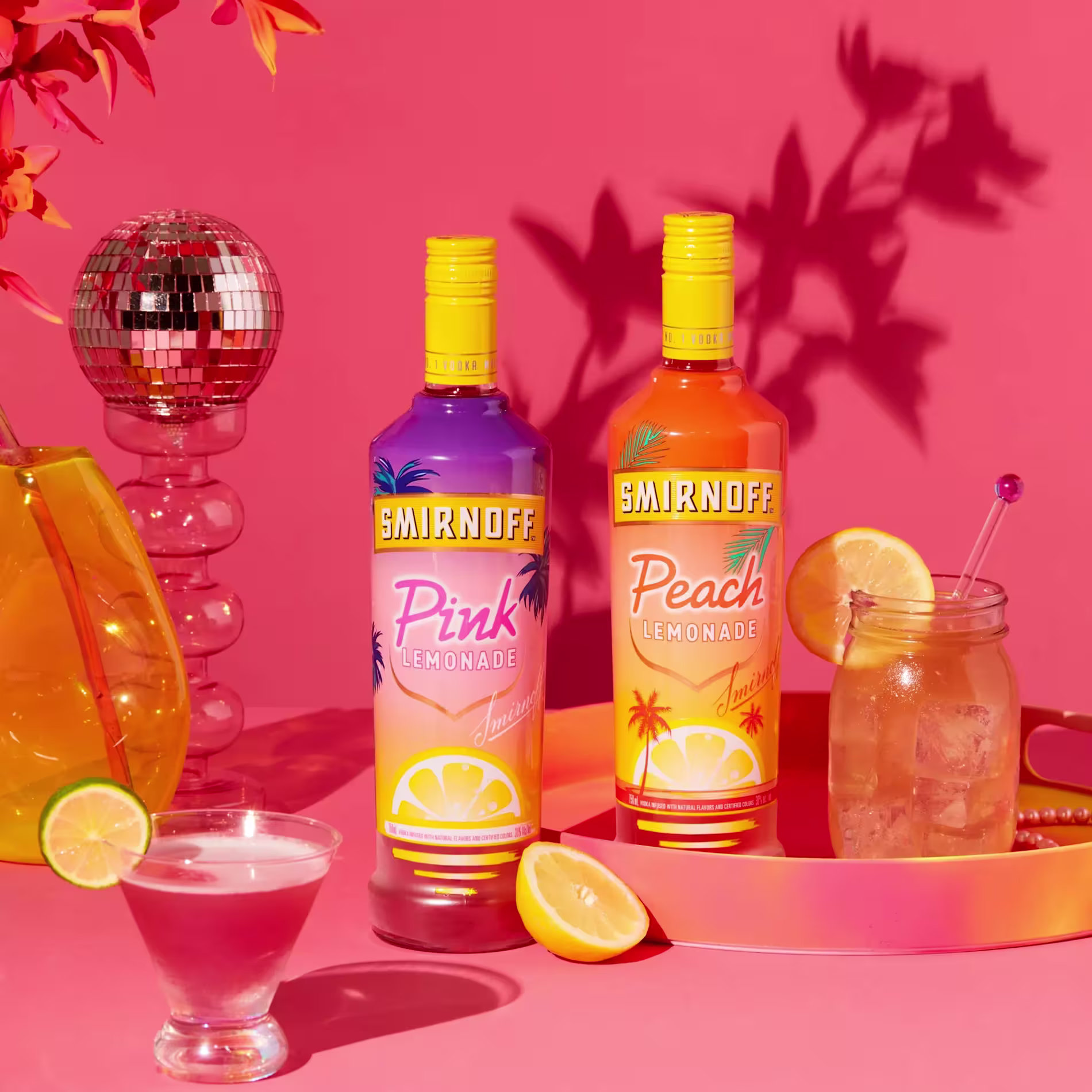 Two bottles of Smirnoff Lemonade, Pink and Peach flavors, stand on a pink surface with cocktails, a lemon slice, a disco ball, and a vase with orange flowers.
