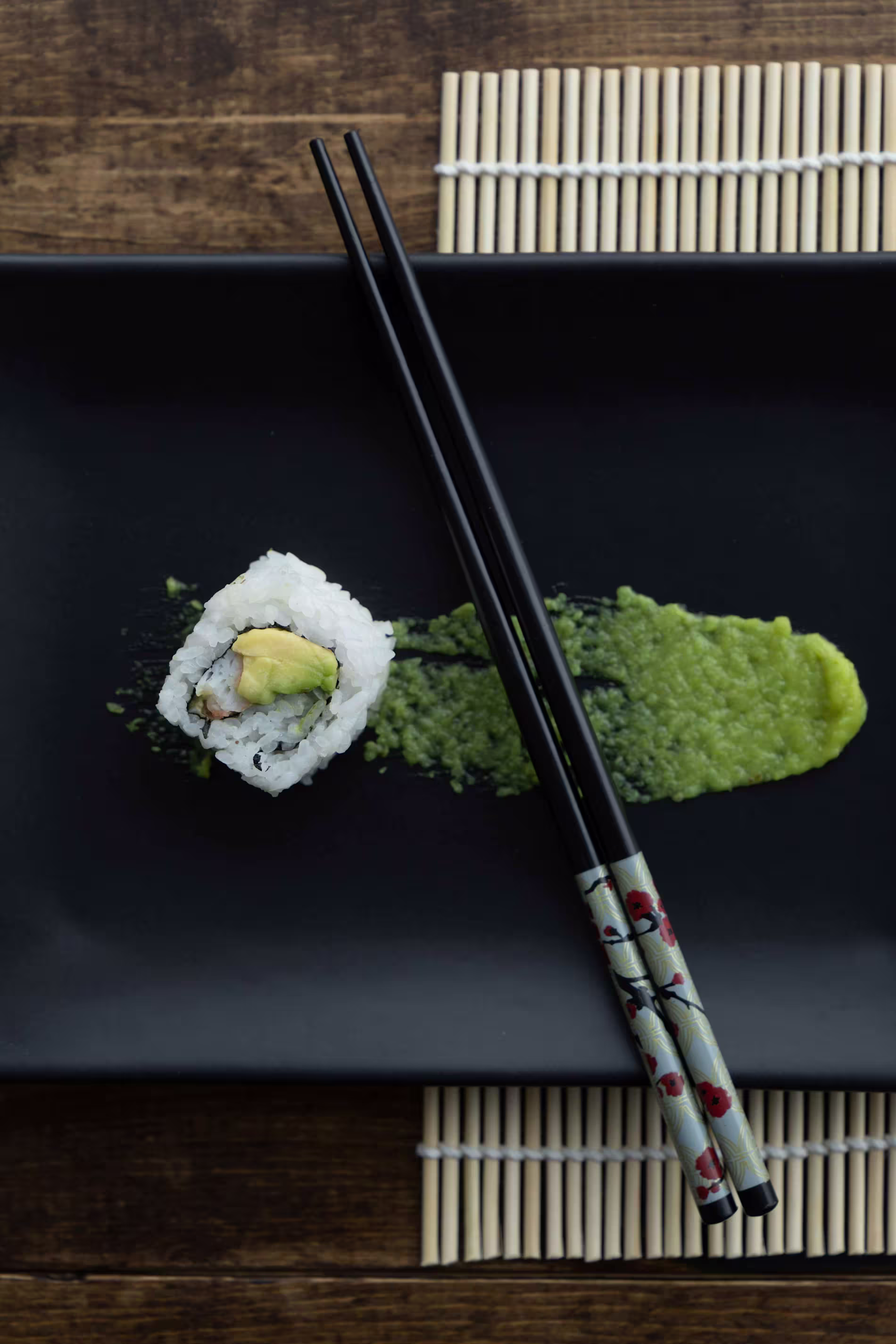 A single sushi roll with avocado and rice is placed on a black plate with a streak of green wasabi, accompanied by a pair of black chopsticks.