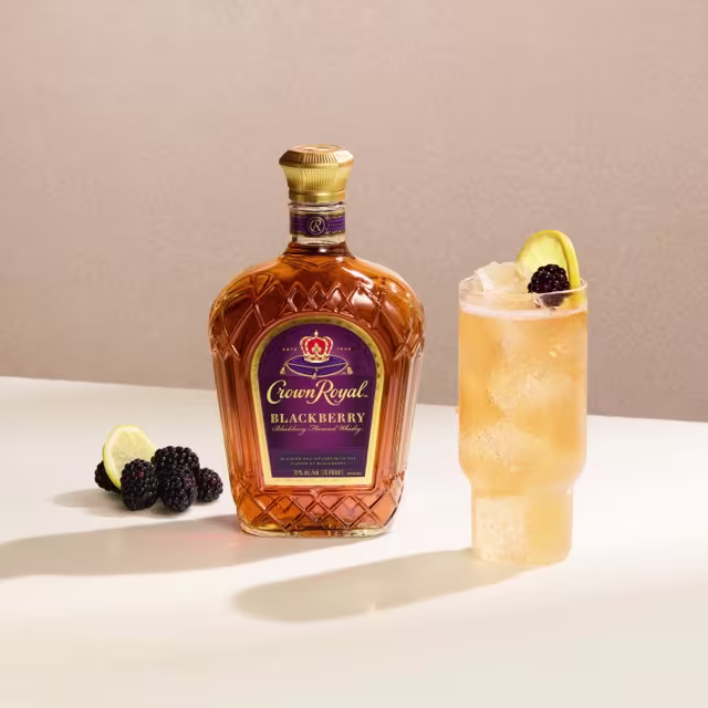A bottle of Crown Royal Blackberry whisky beside a cocktail garnished with a blackberry and lemon slice, with more blackberries and lemon slices on the table.