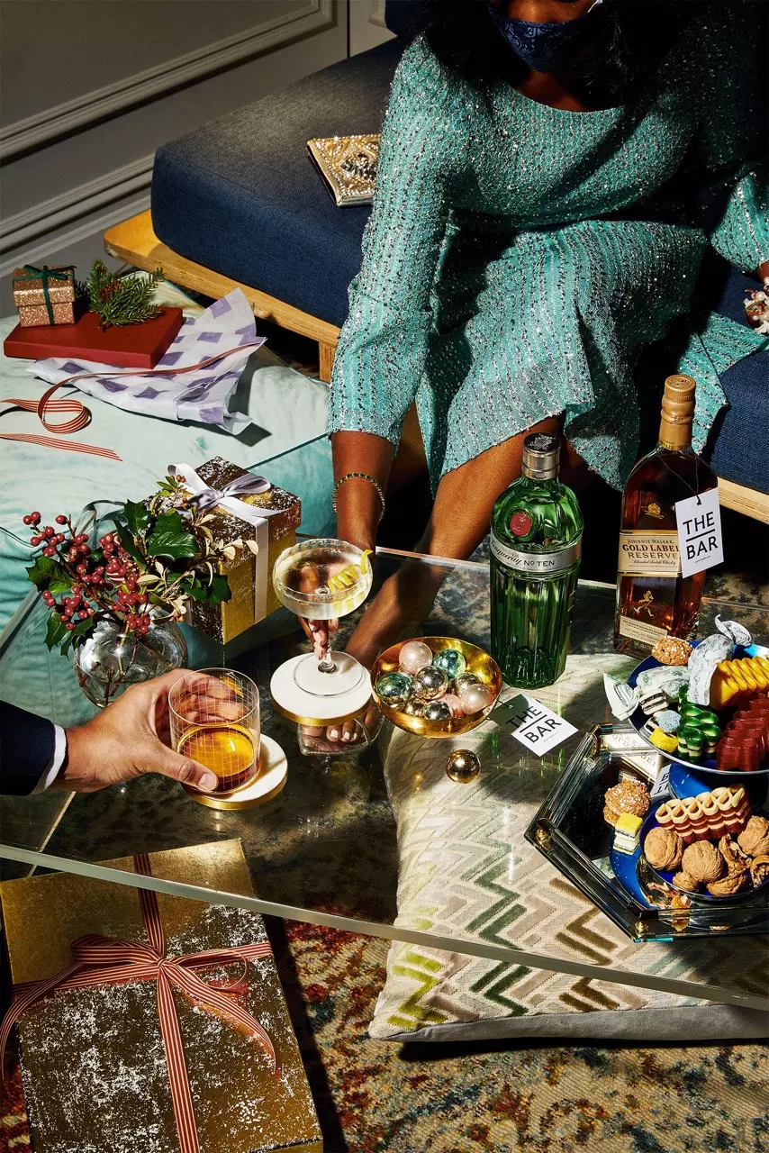 A glass coffee table with drinks, wrapped gifts, holiday decorations, bottles of gin and whiskey, and a tray of assorted cookies; two people are seated nearby.