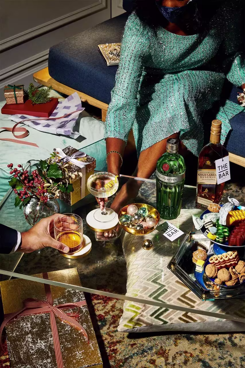 A glass coffee table with drinks, wrapped gifts, holiday decorations, bottles of gin and whiskey, and a tray of assorted cookies; two people are seated nearby.