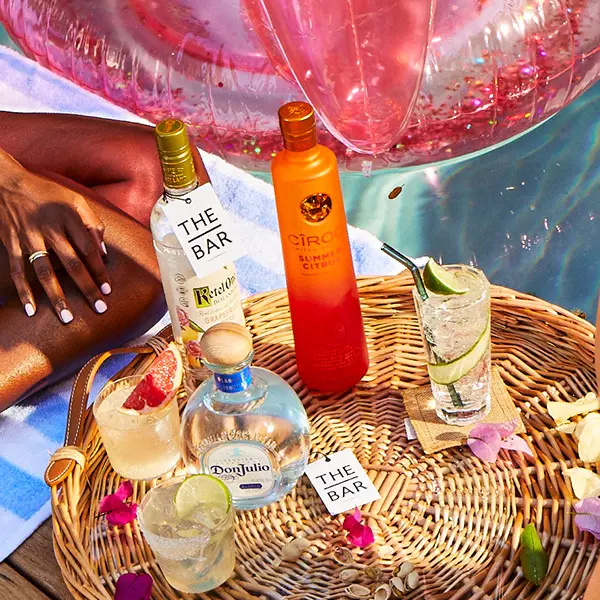 A poolside scene with bottles of tequila and vodka, cocktails with garnishes, a wicker tray, and a pink inflatable float, next to a person sitting on a towel.