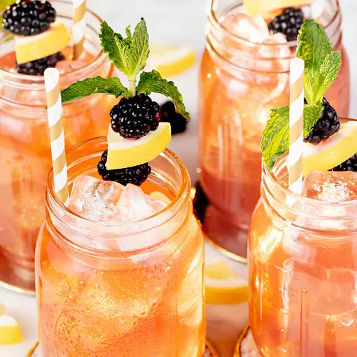 Four mason jar glasses filled with iced pink drinks, garnished with lemon slices, blackberries, and mint sprigs, each with a striped straw.