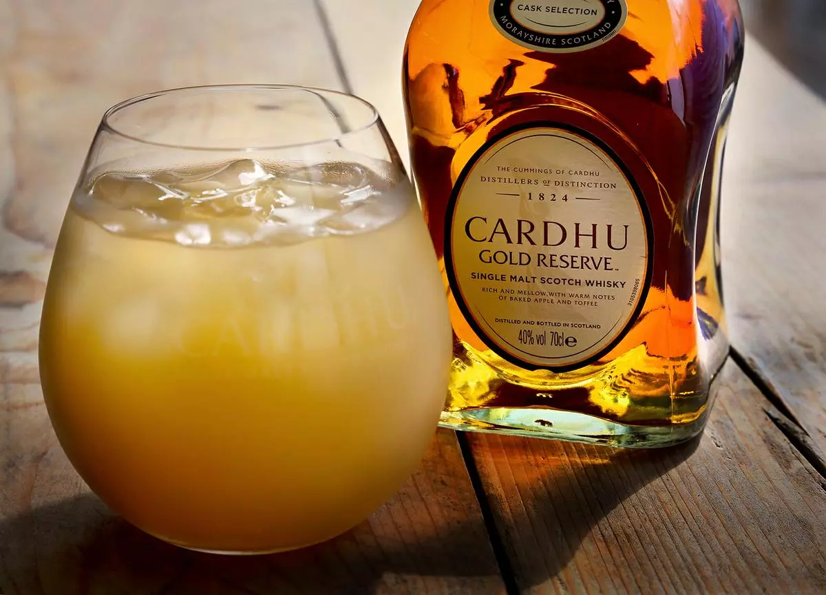A glass of whisky cocktail with ice sits next to a bottle of Cardhu Gold Reserve single malt Scotch whisky on a wooden surface.