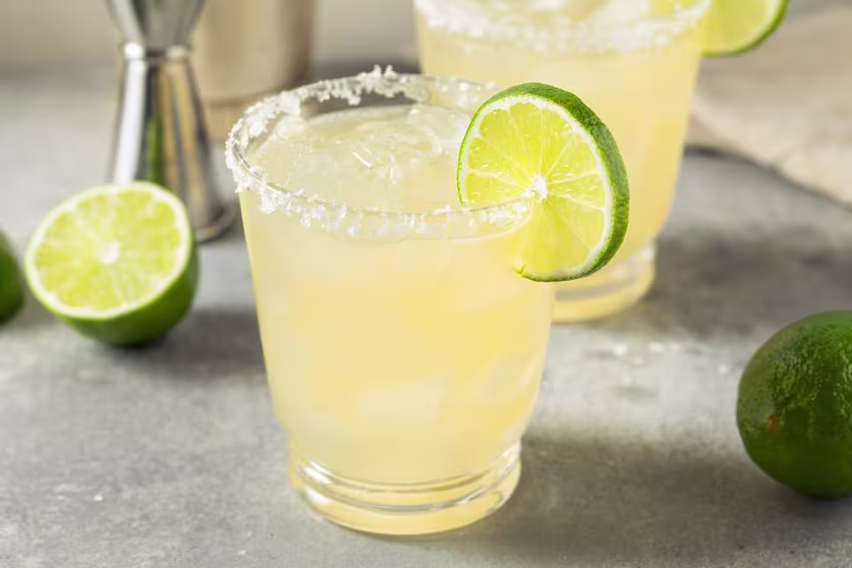 A glass of margarita with ice, a salted rim, and a lime wheel garnish sits on a gray surface with whole and sliced limes nearby.
