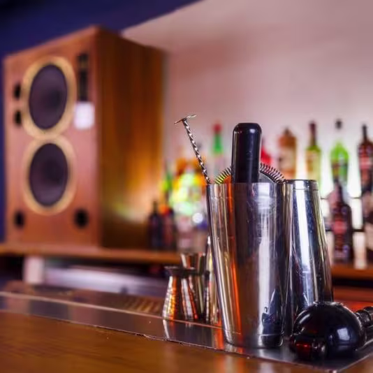 La foto muestra un primer plano de una barra con varias herramientas de bar como una coctelera y un colador en foco. En el fondo, hay una variedad de botellas desenfocadas, con diferentes tipos de alcohol, y un altavoz grande en el lado izquierdo.
