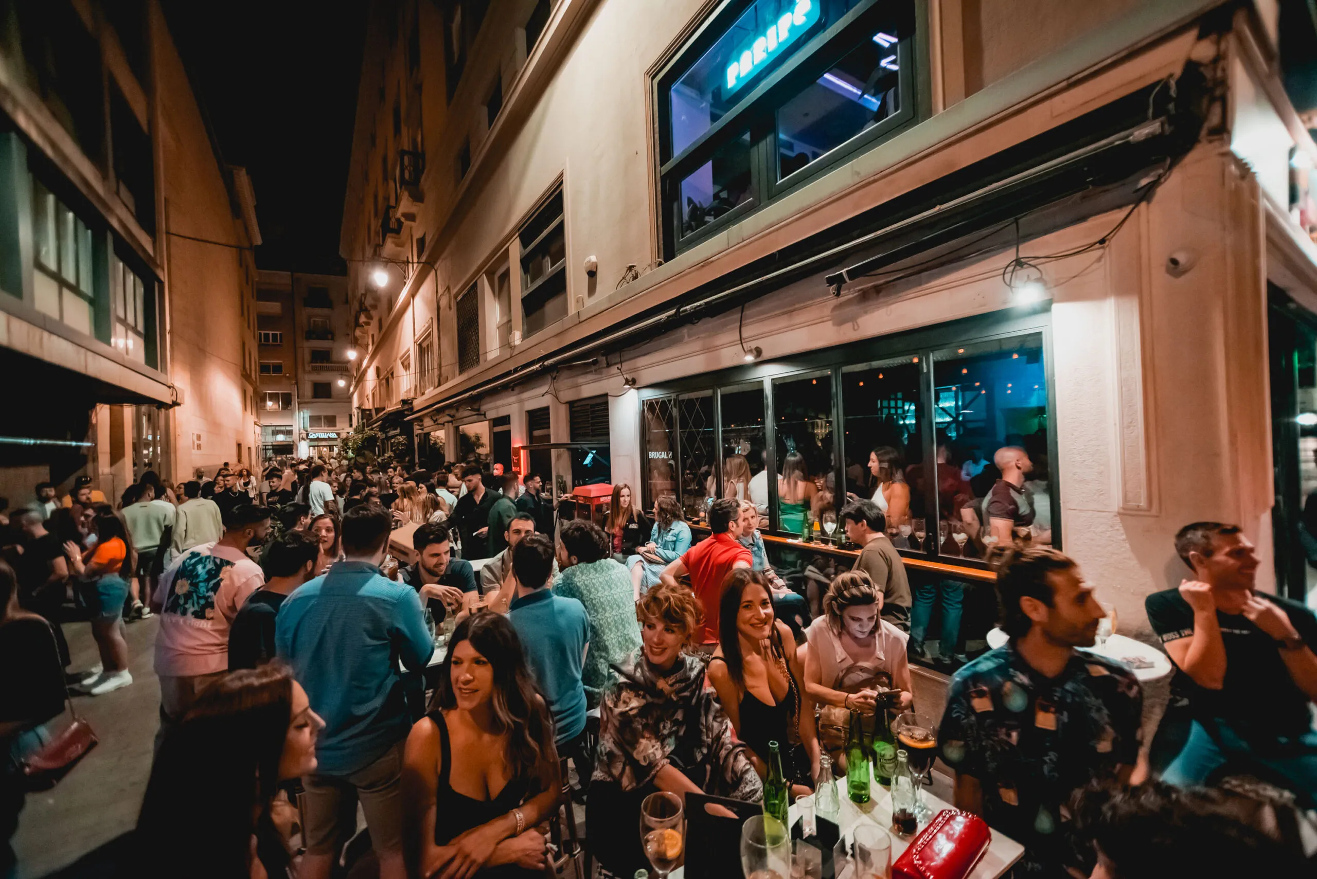 La imagen muestra una vibrante calle nocturna frente al bar Paripé. Grupos de personas disfrutan al aire libre entre risas, bebidas y buena música.