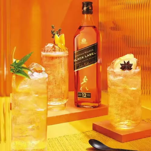 A bottle of Johnnie Walker Black Label whisky is displayed behind three glasses of cocktails, each garnished with herbs and fruit, against an orange background.