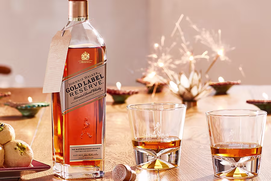 A bottle of Johnnie Walker Gold Label Reserve whisky and two glasses of whisky are on a wooden table with decorative lights, sweets, and tealight candles.