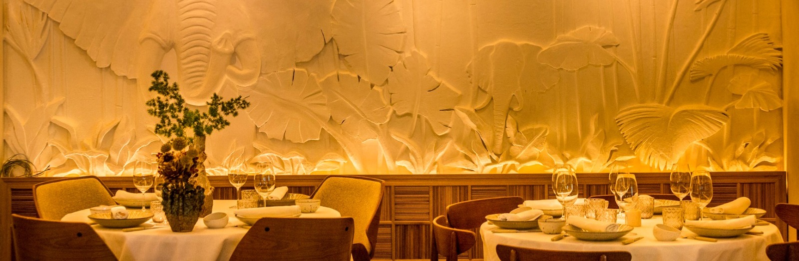Interior de Aarde con una pared decorada con relieves de un elefante y motivos tropicales, mesas elegantemente vestidas y una iluminación cálida.