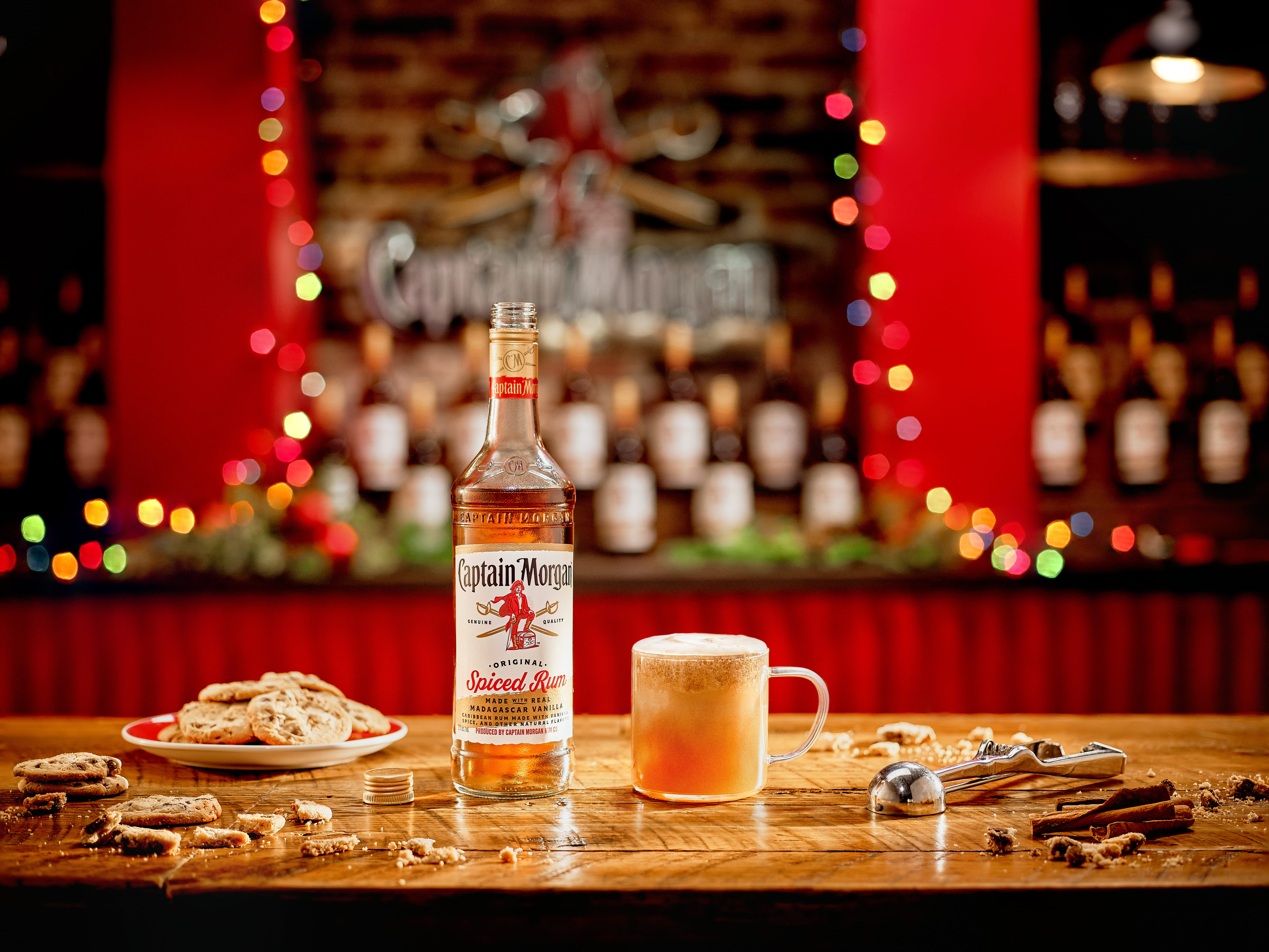 A bottle of Captain Morgan Spiced Rum, a glass of frothy drink, cookies, cinnamon sticks, and an ice scoop on a wooden bar with festive lights in the background.