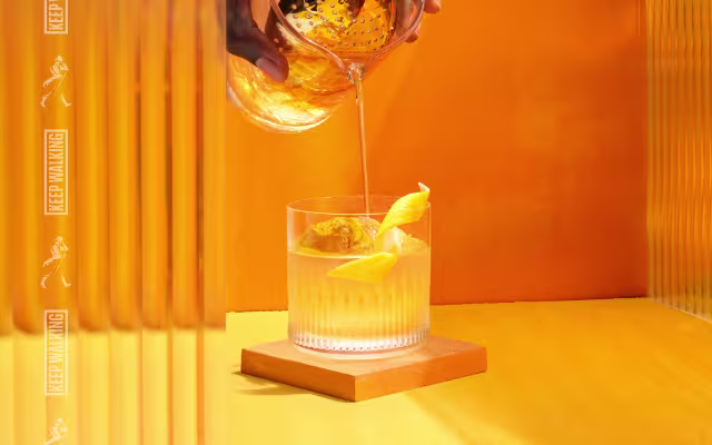 A hand pours an amber-colored drink through a strainer into a glass with ice and a lemon twist, set against a bright orange background.
