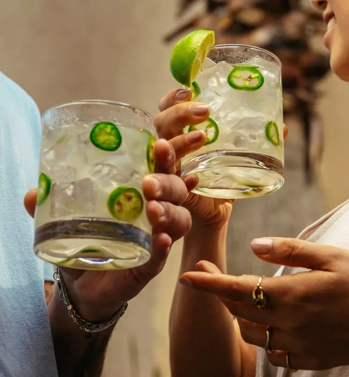 Two people holding glasses of a clear cocktail with ice, lime wedges, and sliced jalapeños.