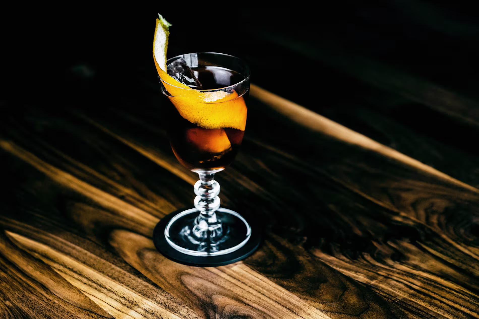 A cocktail in a stemmed glass with ice and an orange peel garnish, placed on a dark wooden table.
