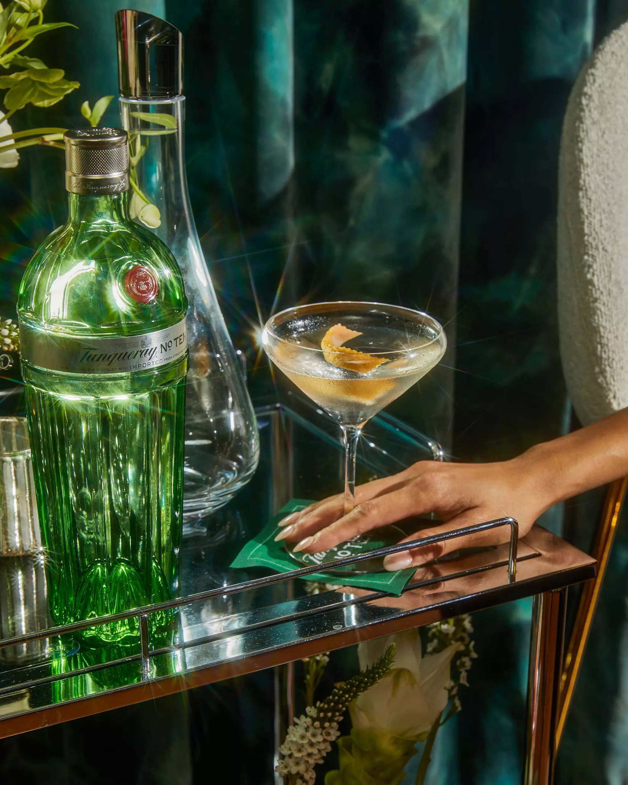 A hand rests on a bar cart next to a green Tanqueray No. Ten gin bottle, a martini glass with a citrus twist, and a clear decanter.