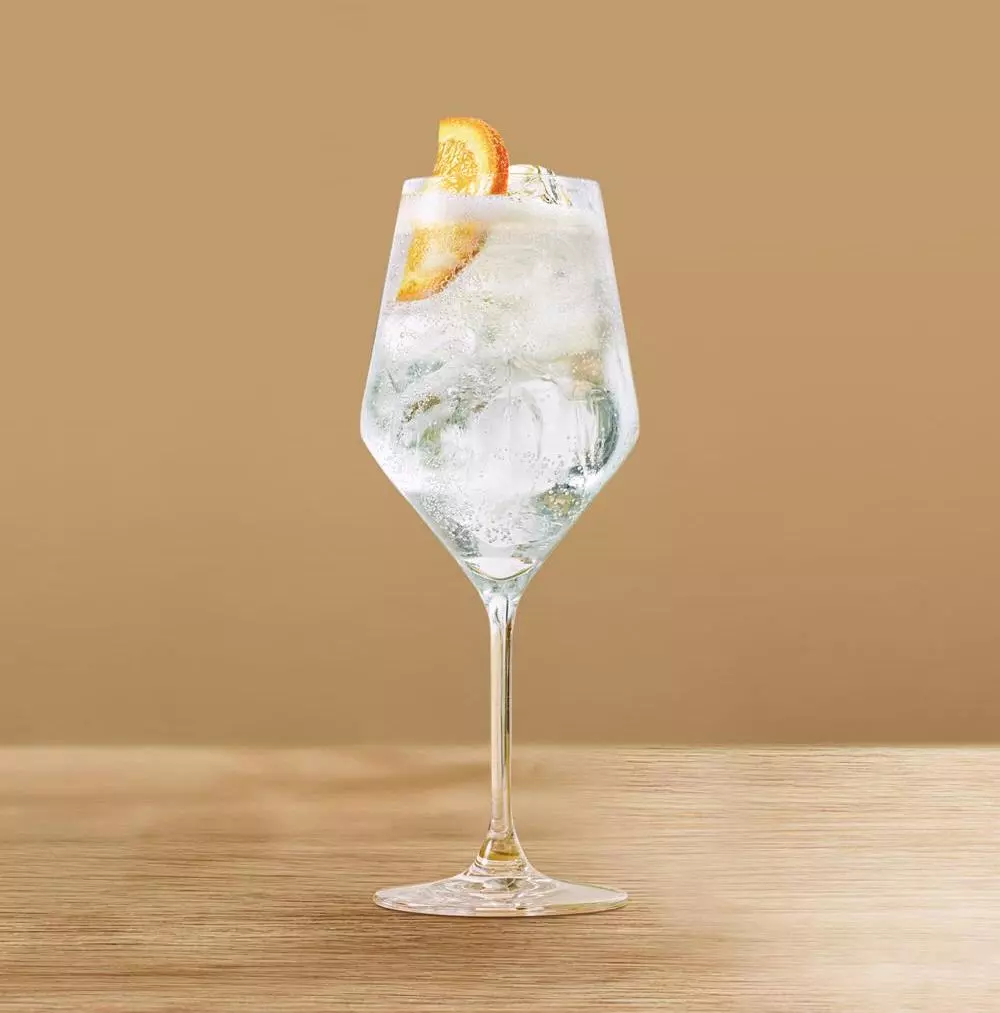 A wine glass filled with a clear sparkling drink, ice cubes, and an orange slice, placed on a wooden surface against a beige background.