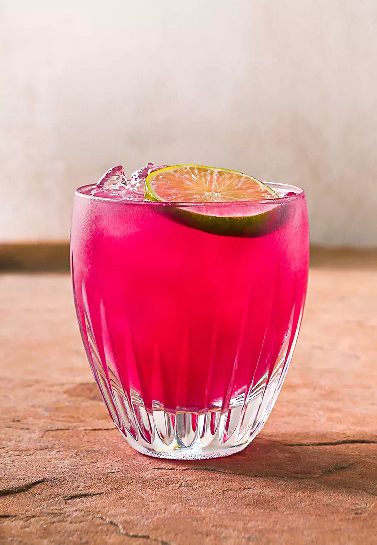 A clear glass filled with bright pink iced beverage, garnished with a slice of lime, sits on a textured brown surface.