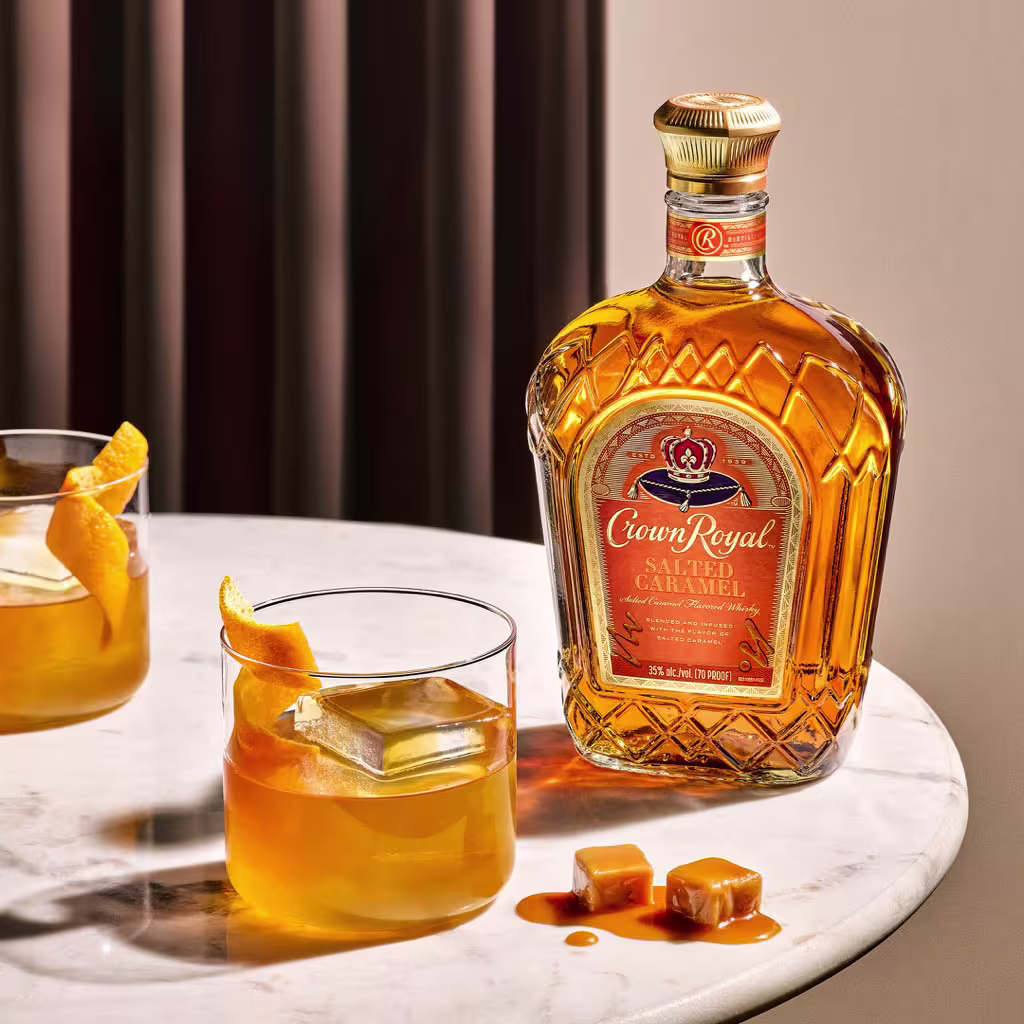 A bottle of Crown Royal Salted Caramel whisky sits on a marble table next to two glasses of whisky with ice and orange peel, and two caramel candies.