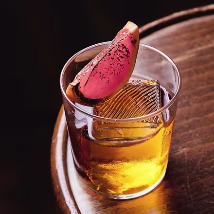 Cóctel en vaso bajo con hielo grande decorado con rodaja de fruta cítrica caramelizada, sobre mesa de madera con luz tenue.