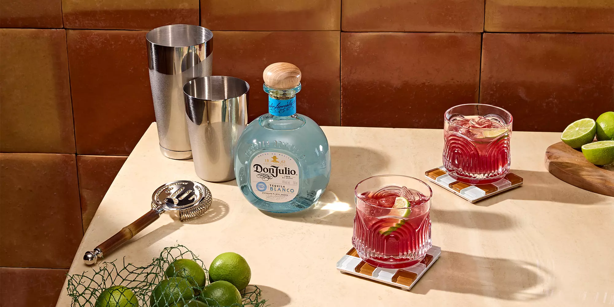 A bottle of Don Julio tequila, two cocktails with lime wedges, a cocktail shaker set, a strainer, whole limes, and lime halves on a beige countertop.