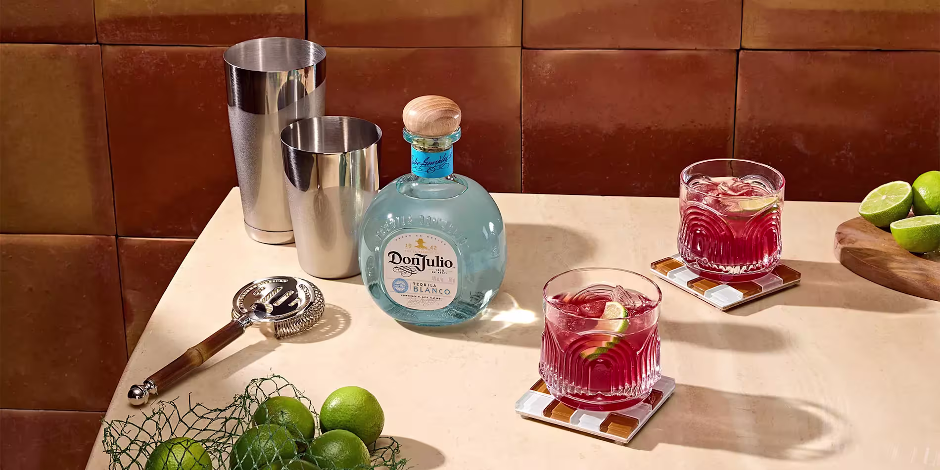 A bottle of Don Julio tequila, two cocktails with lime wedges, a cocktail shaker set, a strainer, whole limes, and lime halves on a beige countertop.