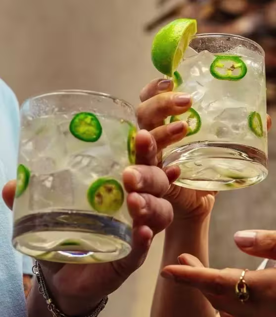 Two hands hold up glasses of a clear iced drink garnished with lime slices and jalapeño rounds.