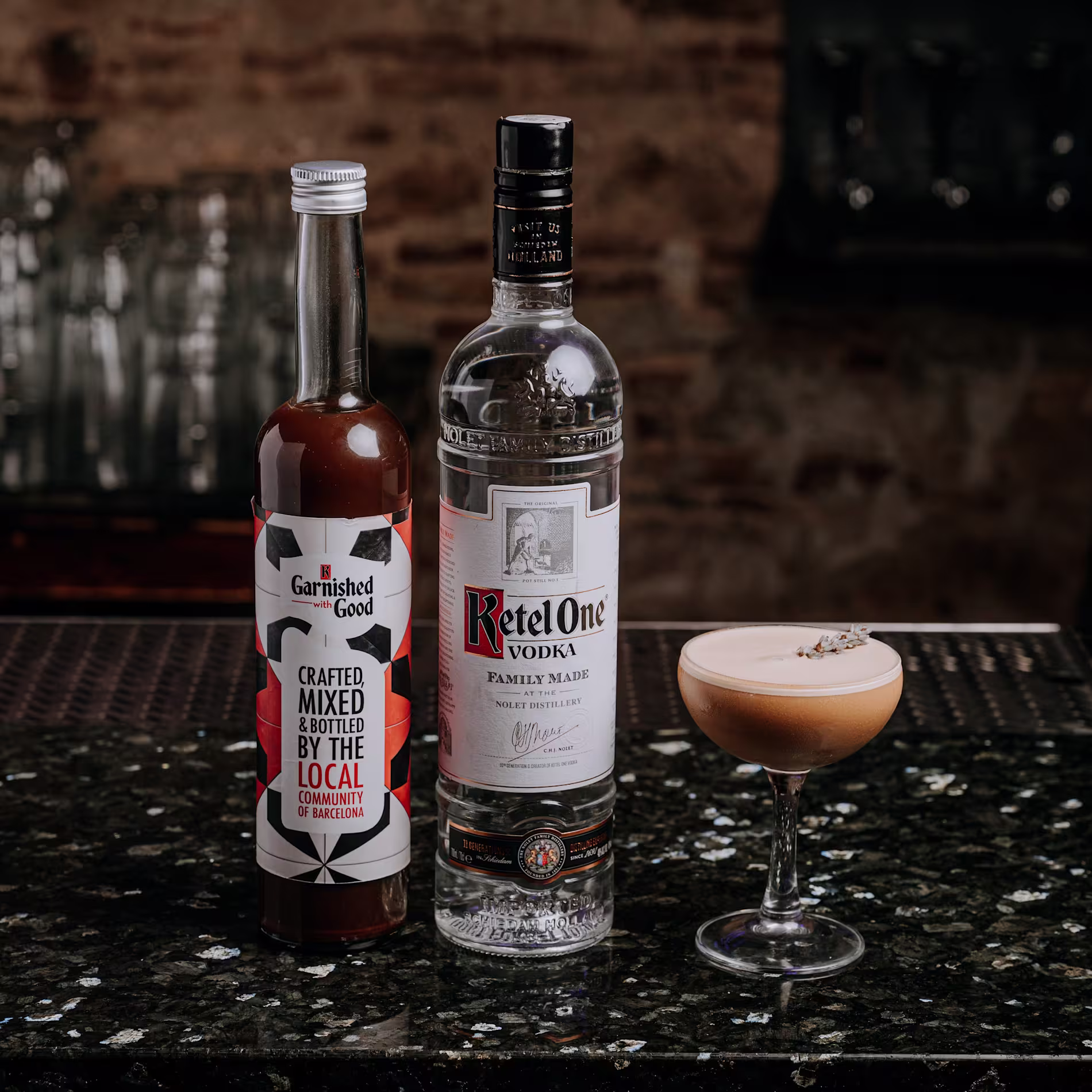 Cóctel con espuma servido en copa junto a botellas de Ketel One y mezcla artesanal sobre una barra negra brillante. Al fondo, estantería con vasos y pared de ladrillo.