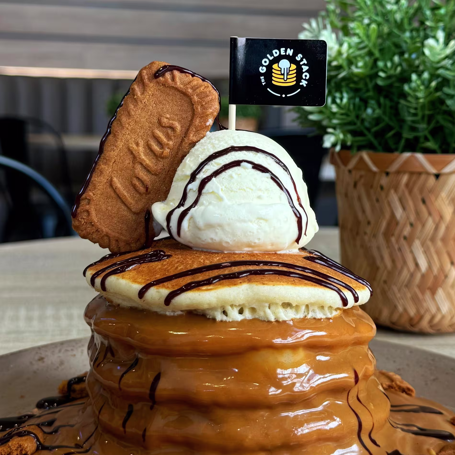 Torre de pancakes cubiertos con crema de Lotus Biscoff, galleta, helado de vainilla y sirope de chocolate, con una banderita del local, sobre fondo de madera clara con el lema "Best pancakes in town".