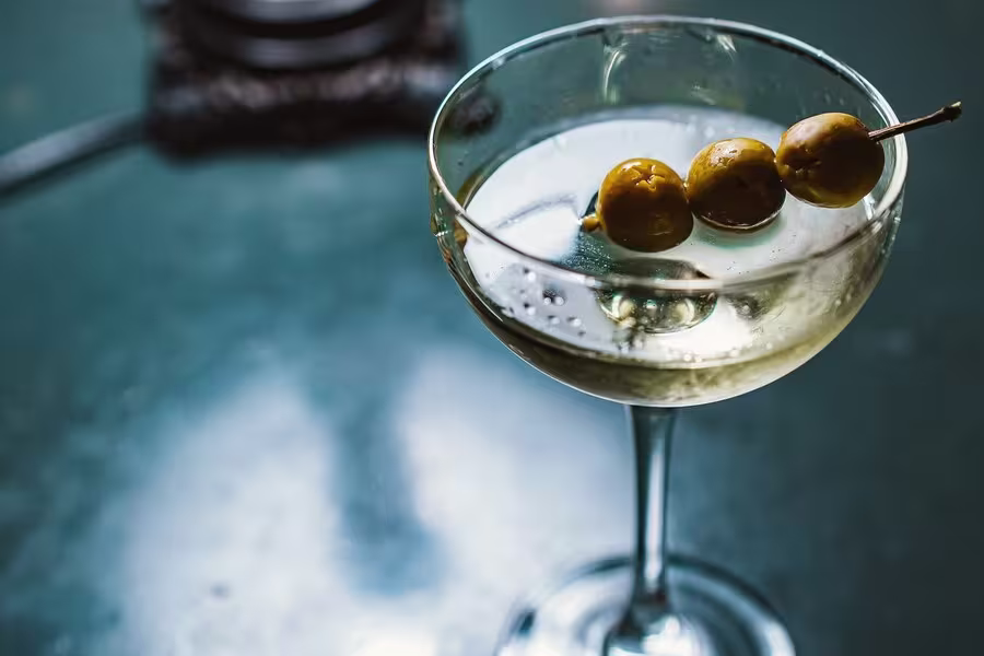 A close-up of a martini in a cocktail glass, garnished with three green olives on a toothpick, on a dark green surface.