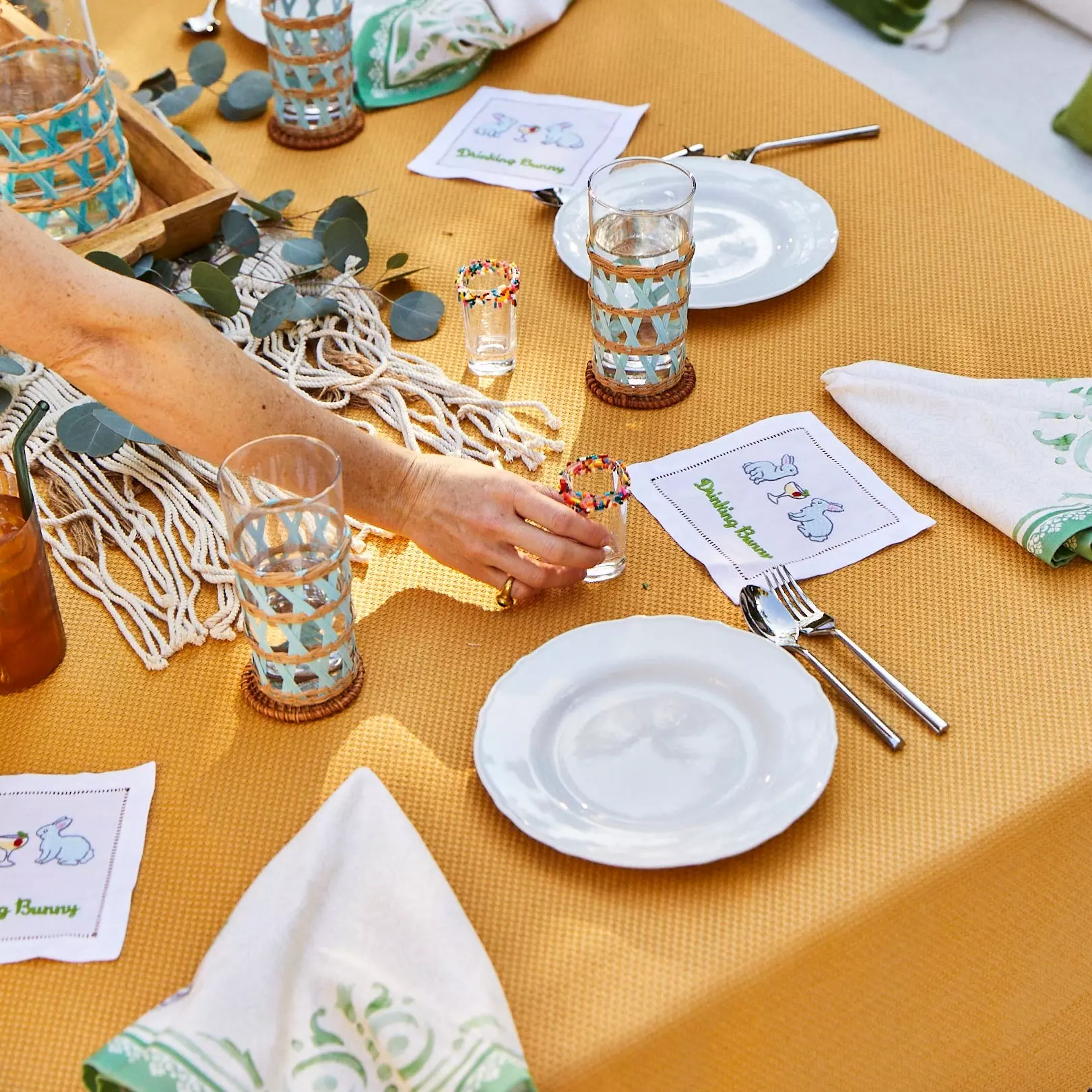 A person sets a table with white plates, patterned napkins, drinking glasses, and “Bunny Bunny” embroidered napkins on a yellow tablecloth.
