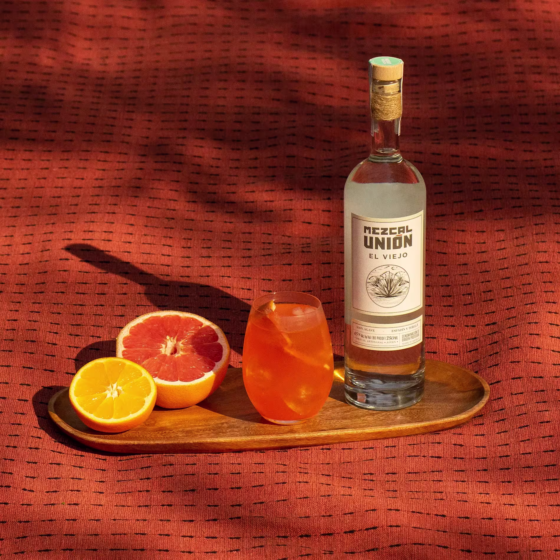 A bottle of Mezcal Unión El Viejo, a cocktail, a halved grapefruit, and a halved orange are arranged on a wooden tray over a textured orange fabric.