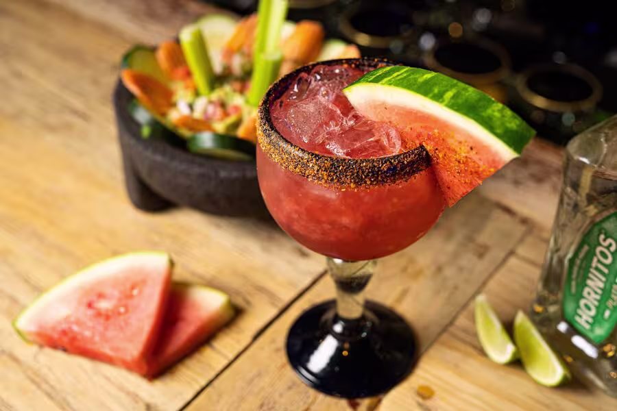 A watermelon cocktail in a glass with a chili rim, garnished with a watermelon slice, sits on a wooden table near lime wedges, a bottle of Hornitos tequila, and a bowl of snacks.