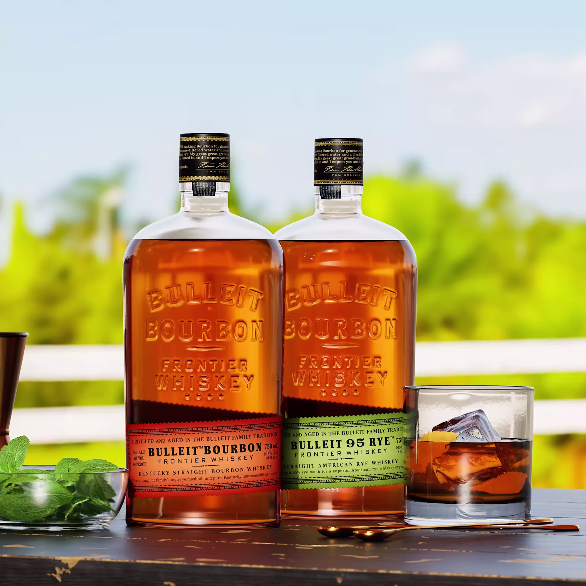 Two bottles of Bulleit whiskey, one bourbon and one rye, sit on an outdoor table next to a glass of whiskey with ice, a sprig of mint, and a blurred green background.