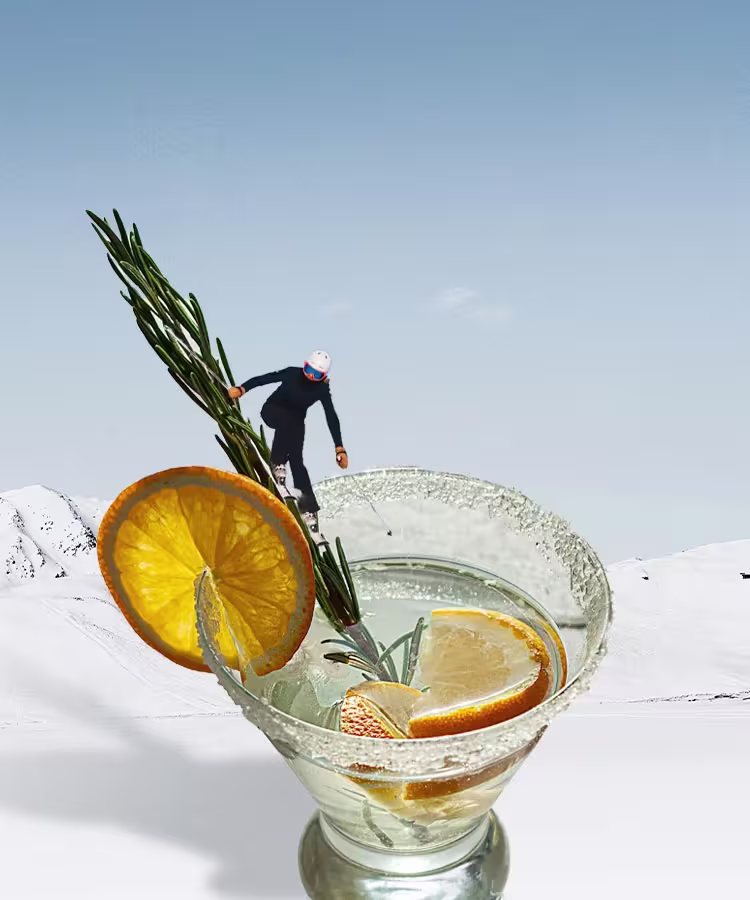 A person skis down a snowy slope that blends into a cocktail glass garnished with lemon slices and rosemary against a clear sky.