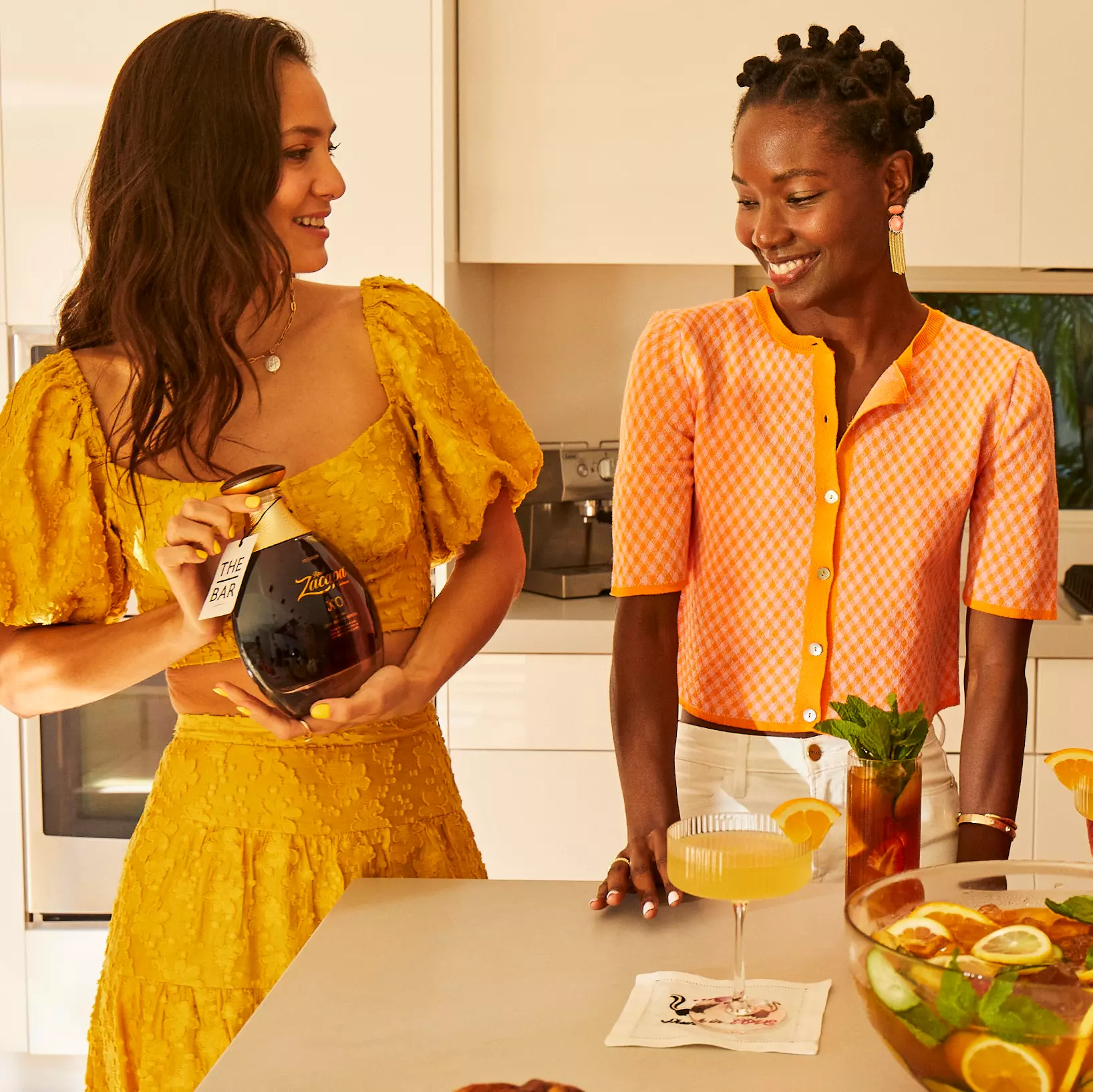 Two women stand in a kitchen; one holds a bottle while the other rests her hand on the counter near a cocktail and a bowl of fruit punch.