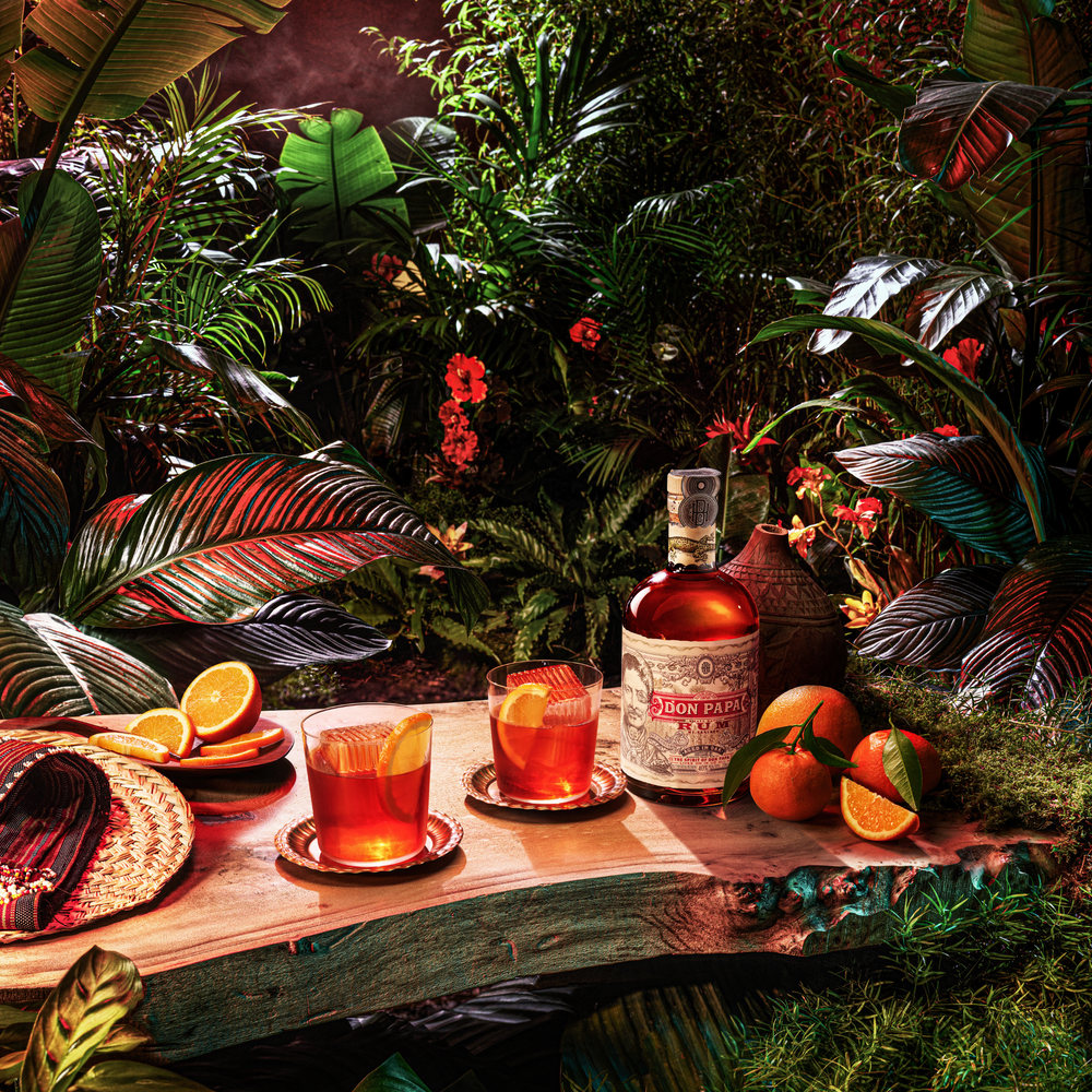 Don Papa rum bottle with two cocktails garnished with orange slices on a wooden table surrounded by tropical plants