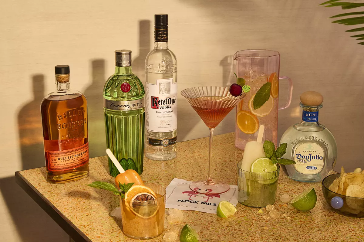 A tabletop with bottles of bourbon, gin, vodka, and tequila, mixed cocktails, a pitcher with fruit, lime wedges, chips, and a napkin with “Flock Tails” printed on it.