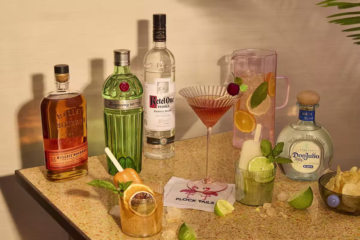 A tabletop with bottles of bourbon, gin, vodka, and tequila, mixed cocktails, a pitcher with fruit, lime wedges, chips, and a napkin with “Flock Tails” printed on it.