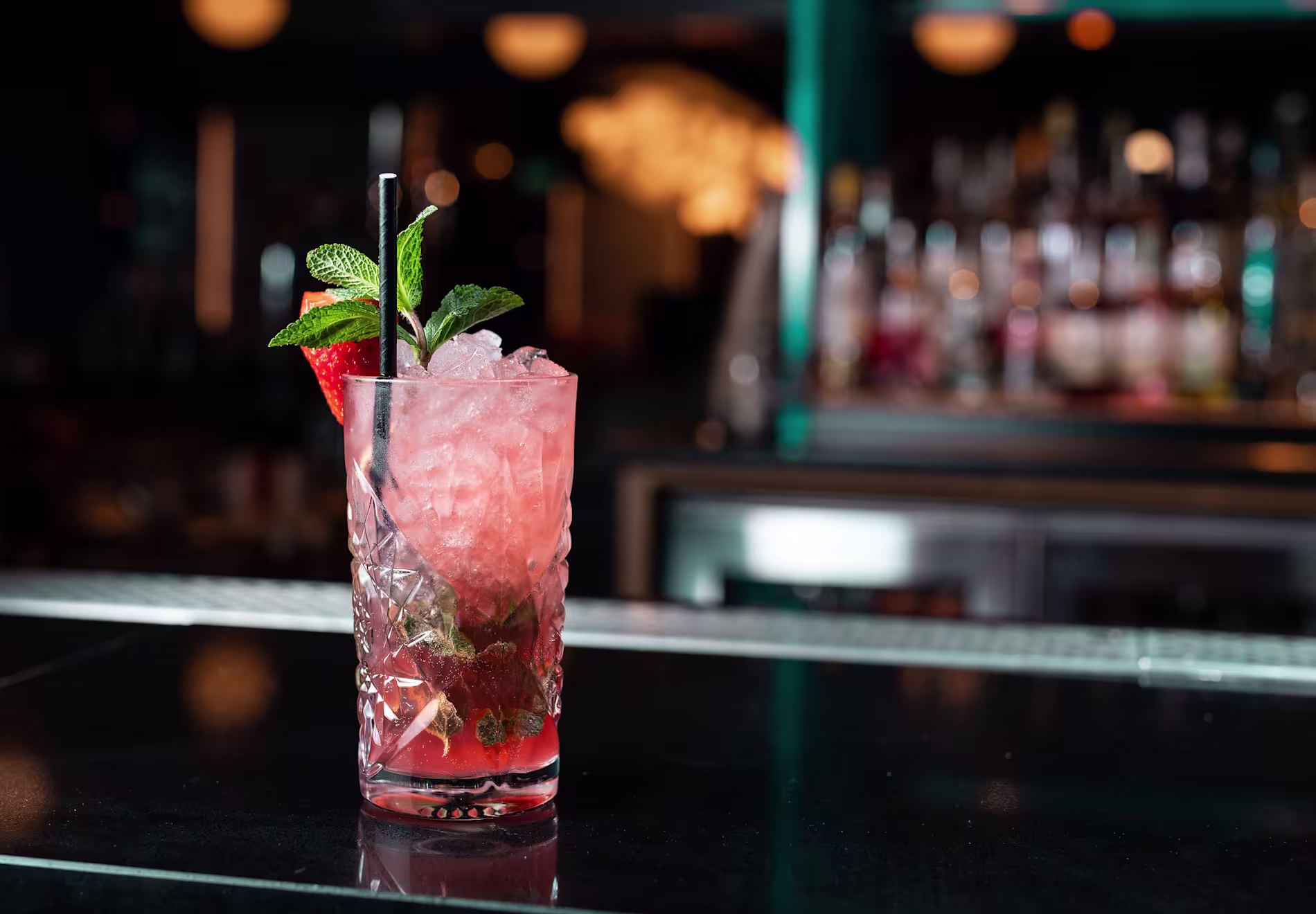 A pink cocktail with crushed ice, mint leaves, and a strawberry garnish in a tall glass sits on a dark bar counter with a blurred background.