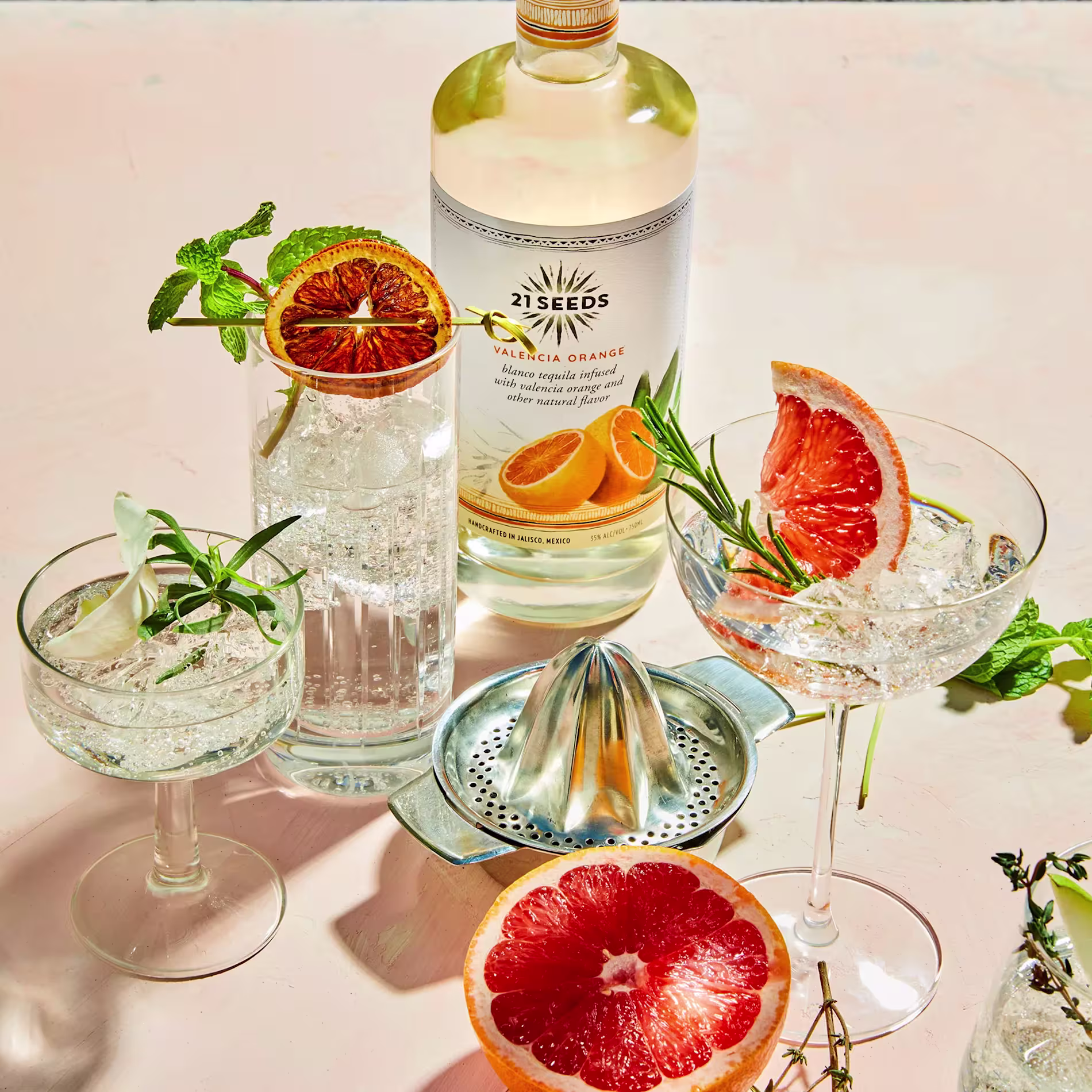 A bottle of 21 Seeds Valencia Orange tequila is surrounded by three cocktails garnished with citrus, herbs, and a halved grapefruit on a light surface.