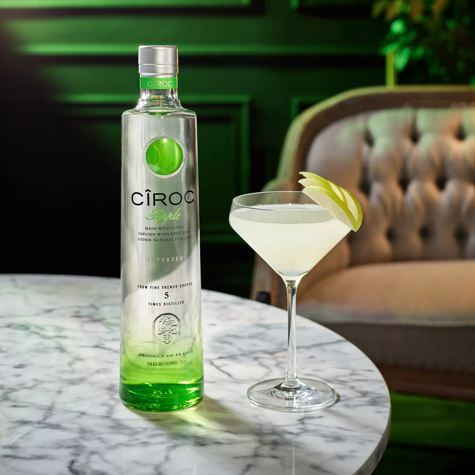A bottle of Ciroc Apple vodka and a martini glass with a light green cocktail garnished with apple slices on a marble table.