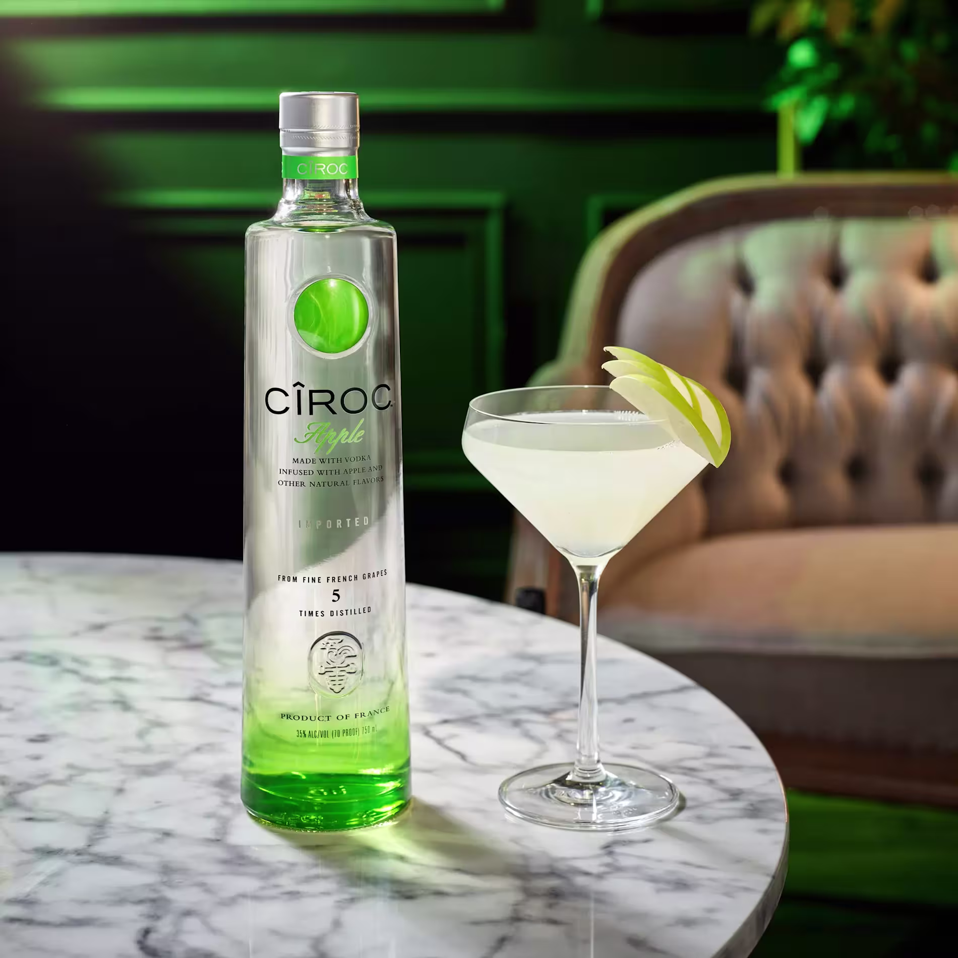 A bottle of Ciroc Apple vodka and a martini glass with a light green cocktail garnished with apple slices on a marble table.