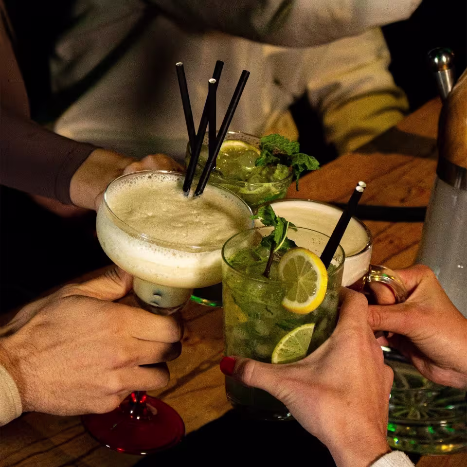 Brindis entre varias personas con cócteles decorados con menta y rodajas de limón sobre una mesa de madera.