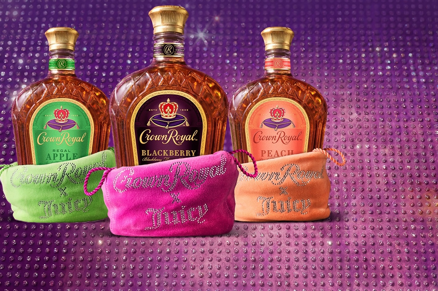 Three Crown Royal flavored whisky bottles, Regal Apple, Blackberry, and Peach, displayed against a sparkling purple backdrop. In front of each bottle is a matching Crown Royal x Juicy Couture velvet bag in green, pink, and peach, embellished with rhinestone lettering.