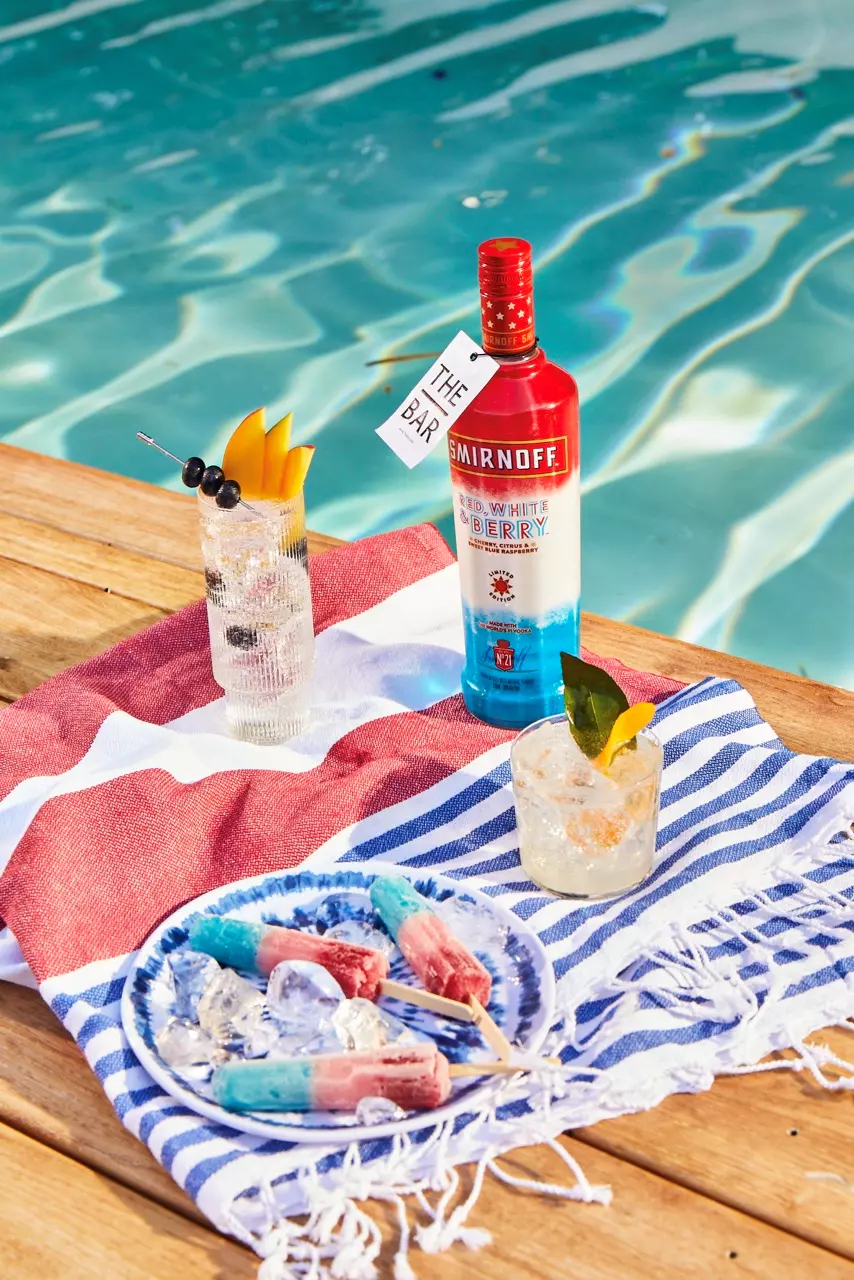A bottle of Smirnoff Red, White & Berry vodka, three cocktails, and popsicles on a towel by a swimming pool.