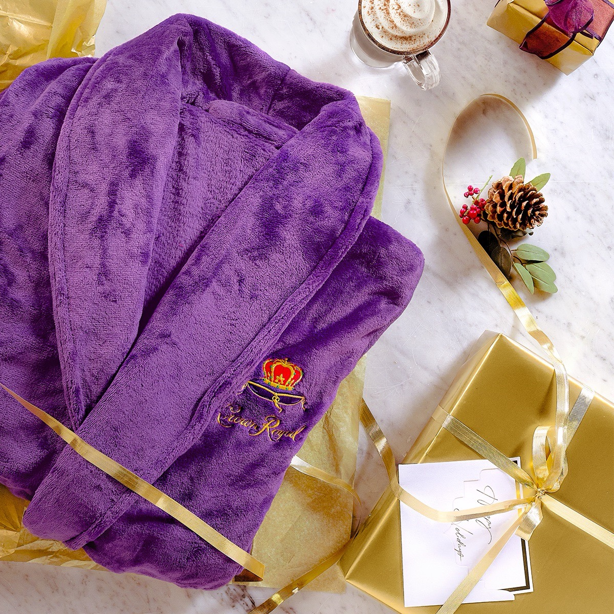 A purple robe with gold embroidery is laid out on yellow tissue paper next to gold-wrapped gifts, a pinecone decoration, and a cup of coffee on a marble surface.