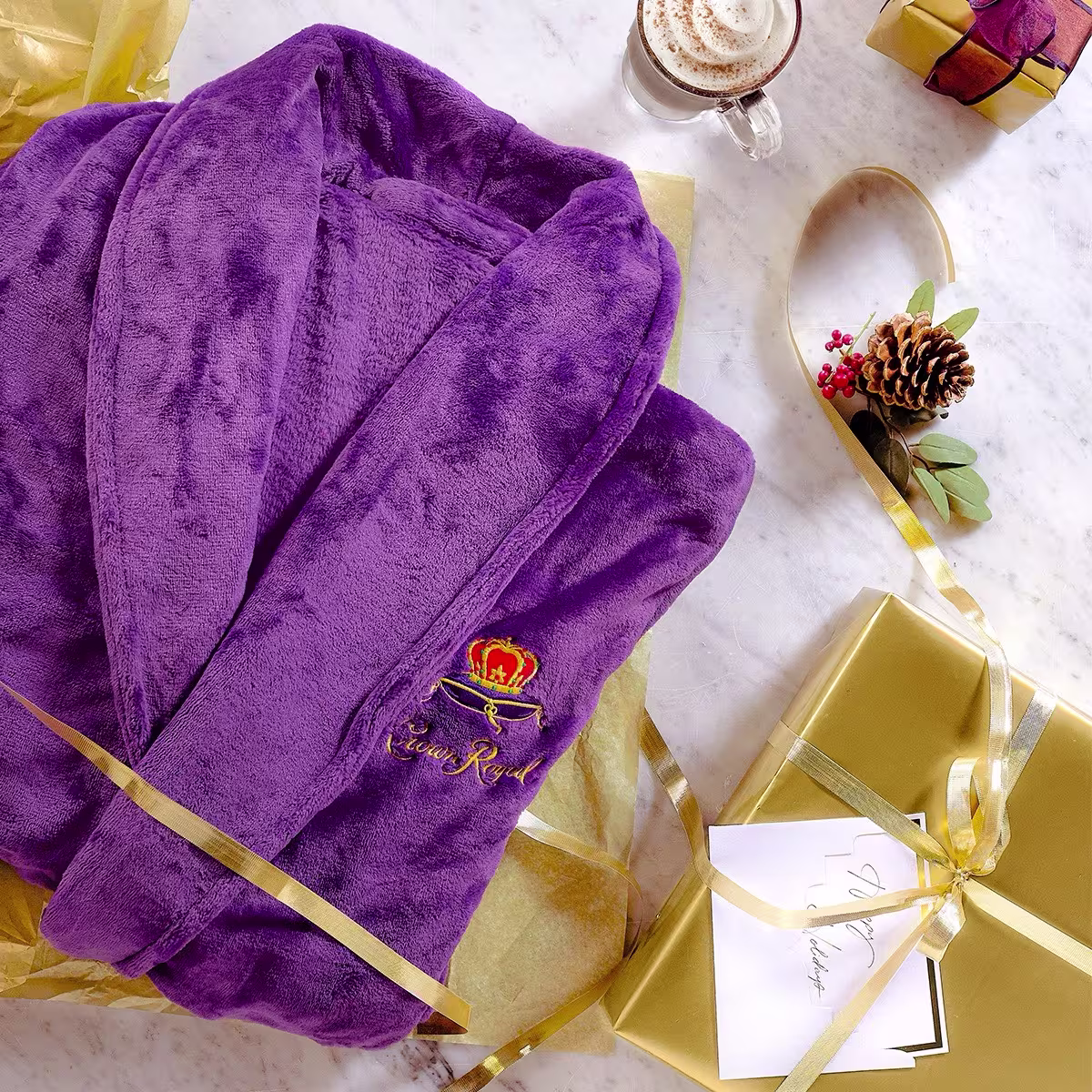 A purple robe with gold embroidery is laid out on yellow tissue paper next to gold-wrapped gifts, a pinecone decoration, and a cup of coffee on a marble surface.